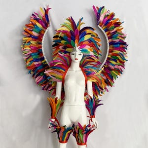 Op de afbeelding: Een mannequin toont een levendig, veelkleurig veren kostuum. Het kostuum omvat een gevederde hoofdtooi, een schouderstuk, manchetknopen en beenbedekkingen. Grote, gebogen veren vleugels met een zilveren rand zijn aan de achterkant bevestigd. De veren zijn in tinten rood, oranje, geel, groen, blauw en paars.