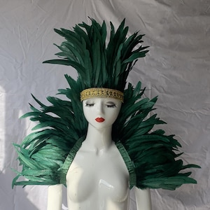 Peut inclure: Un mannequin est orné d'une coiffe de plumes vertes et d'une pièce d'épaule assortie. Un bandeau doré avec des détails complexes entoure la tête. Le mannequin porte du rouge à lèvres rouge.