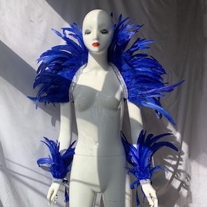 May include: A mannequin showcases a blue feather shoulder piece, arm cuffs, and a rhinestone-studded strap. The vivid blue feathers create a dramatic effect. The cuffs are adorned with matching feathers and rhinestones.