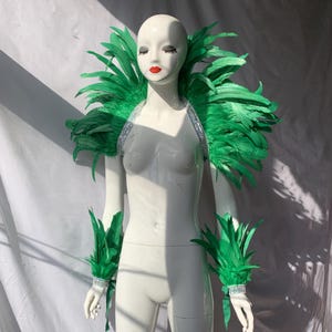 May include: A mannequin showcases a green feather shoulder piece and matching wrist cuffs. The shoulder piece features a silver strap with rhinestones. The wrist cuffs are also adorned with rhinestones. The feathers are a vibrant green colour.