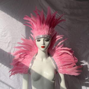 May include: A pink feather headdress and shoulder piece set on a mannequin. The headdress has a jeweled headband and tall feathers. The shoulder piece has a jeweled strap and large feathers. The mannequin has red lipstick.
