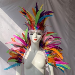 May include: A flamboyant, multi-coloured feather headdress and shoulder piece. The costume features a rhinestone-studded headband and straps, with a dazzling array of rainbow-coloured feathers. The costume is displayed on a mannequin.
