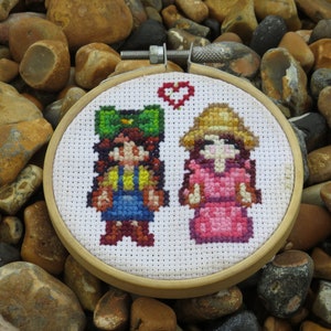 May include: Cross-stitch embroidery hoop featuring two figures of people. The figures are a girl with a green hat and a girl with a straw hat. The background is white with a red heart.