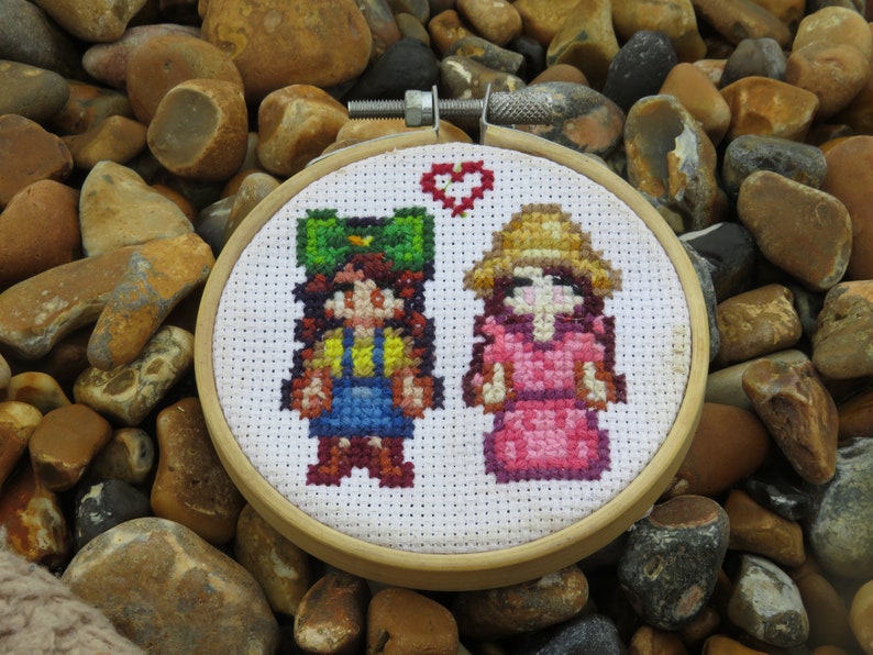 May include: Cross-stitch embroidery hoop featuring two figures of people. The figures are a girl with a green hat and a girl with a straw hat. The background is white with a red heart.