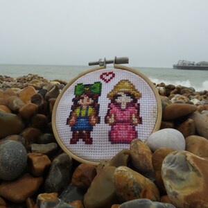 May include: A white cross-stitch embroidery hoop with a heart design. The embroidery features two pixelated figures, one in a green hat and blue overalls, and the other in a pink dress and a straw hat.