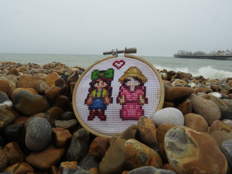May include: A white cross-stitch embroidery hoop with a heart design. The embroidery features two pixelated figures, one in a green hat and blue overalls, and the other in a pink dress and a straw hat.