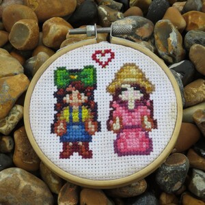 May include: A white embroidery hoop with a cross-stitch design of two figures. The figures are a girl with a green hat and a girl with a straw hat. A red heart is stitched above the figures.
