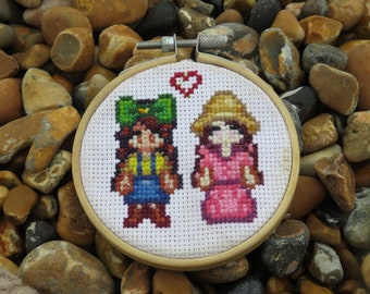 May include: A white embroidery hoop with a cross-stitch design of two figures. The figures are a girl with a green hat and a girl with a straw hat. A red heart is stitched above the figures.