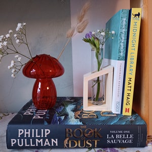 May include: A red glass mushroom-shaped vase with white flowers, a small glass vase with purple flowers in a wooden stand, and three books stacked on top of each other. The books are titled "The Midnight Library", "Before the Coffee Gets Cold", and "The Book of Dust".