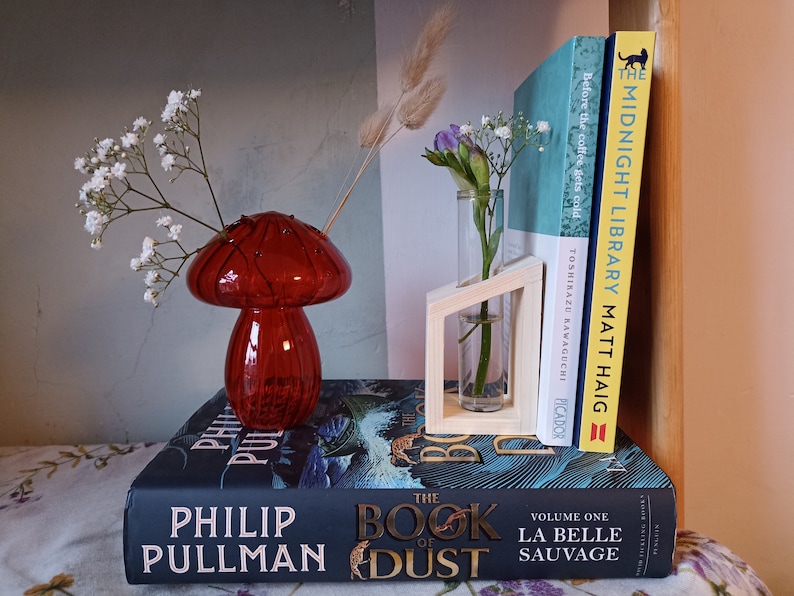 May include: A red glass mushroom-shaped vase with white flowers, a small glass vase with purple flowers in a wooden stand, and three books stacked on top of each other. The books are titled "The Midnight Library", "Before the Coffee Gets Cold", and "The Book of Dust".