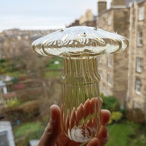 May include: A clear glass mushroom-shaped decorative object with a ribbed stem. The object is held in a hand against a blurry background of buildings and greenery.
