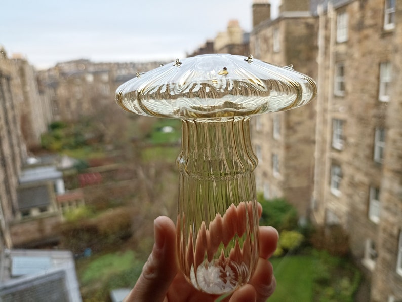 May include: A clear glass mushroom-shaped decorative object with a ribbed stem. The object is held in a hand against a blurry background of buildings and greenery.