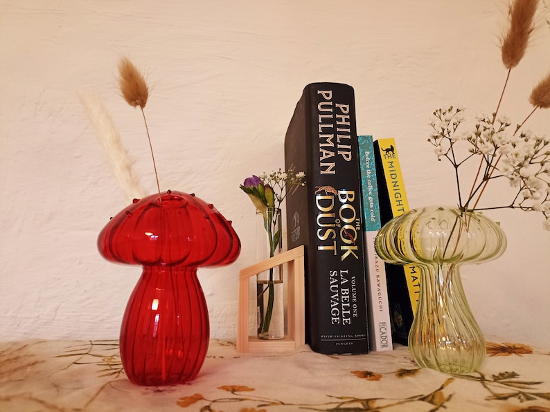 May include: Two glass mushroom-shaped vases, one red and one green, with dried flowers and a stack of books behind them. The books are titled "The Book of Dust" by Philip Pullman and "La Belle Sauvage" by Philip Pullman.