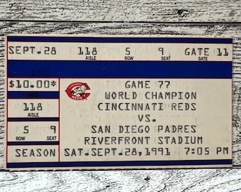 May include: A vintage Cincinnati Reds baseball ticket from a 1991 game against the San Diego Padres at Riverfront Stadium. The ticket is cream-colored with red and blue text and details, including the date, time, and seat information.