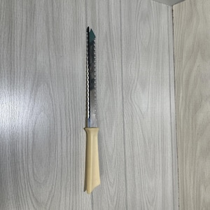 May include: A serrated knife featuring a long, thin, metallic blade with a sharp, jagged edge and a smooth, cream-colored plastic handle. The knife is positioned vertically, showcasing the contrast between the reflective steel and the solid handle.