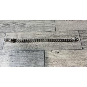 May include: A silver-toned metal bracelet with a rectangular link design. The bracelet is laid flat on a wooden surface. The clasp is visible on the left side. The links are uniform in size and shape.