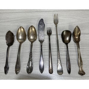 May include: An assortment of antique silverware, including spoons, forks, and a butter knife, arranged on a light gray surface. The silverware has a tarnished, vintage appearance, with ornate handle designs and varying spoon bowl shapes. The collection suggests a formal dining setting.