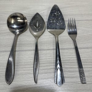 May include: Four vintage silver serving utensils: a soup ladle, a slotted spoon, a pie server, and a fork. The utensils are arranged on a light wood-grain surface. The ladle has a round bowl and a decorative handle. The pie server has a triangular blade.