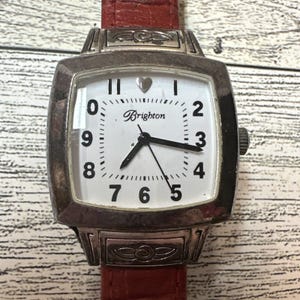 May include: A wristwatch with a square silver-tone metal case and a textured red leather strap. The white dial features black Arabic numerals, black hands, and the word Brighton printed in black. A small silver heart replaces the twelve o'clock marker.