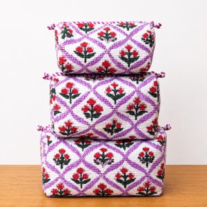 May include: A set of three rectangular fabric storage bins stacked vertically. Each bin features a white background with a repeating purple diamond lattice pattern and a central red floral motif with green leaves on each side of the containers.