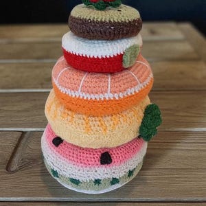 May include: A stack of crocheted food items, including a watermelon, orange, peach, and chocolate donut, topped with a strawberry. The handmade items are in various colors, including pink, orange, red, brown, and green.