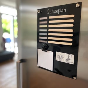 Könnte beinhalten: Schwarzes magnetisches Speiseplan-Board mit dem Wort "Speiseplan" in weißer Schrift. Es verfügt über Schlitze für Holzstäbchen, einen kleinen Notizblock und eine Klammer. Das Board ist an einem Kühlschrank aus Edelstahl befestigt.