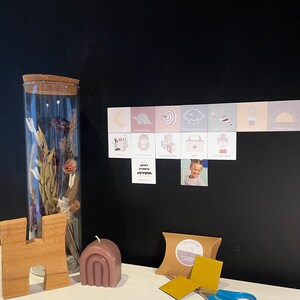 May include: A set of colorful magnetic weekday cards with illustrations and text in German. The cards are displayed on a black chalkboard with a small photo of a child. The set also includes a small brown box with gold magnetic squares and a pair of blue scissors.