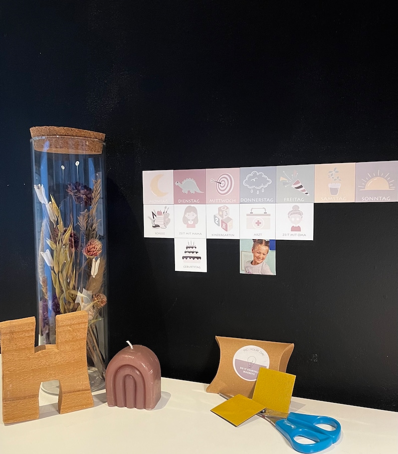 May include: A set of colorful magnetic weekday cards with illustrations and text in German. The cards are displayed on a black chalkboard with a small photo of a child. The set also includes a small brown box with gold magnetic squares and a pair of blue scissors.