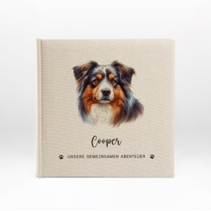 Könnte beinhalten: Ein Album aus beigefarbenem Leinen mit einer Schwarzweißillustration eines Hundes auf der Vorderseite. Der Hund hat braunes, schwarzes und weißes Fell. Das Album trägt den Namen "Cooper" in schwarzer kursivschrift und den Text "UNSERE GEMEINSAMEN ABENTEUER" in schwarzer Schrift unter dem Hund.