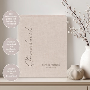 May include: A beige linen-covered Stammbuch, or family book, with the title in elegant script. The book is displayed on a shelf with a white vase holding branches with white buds and a smaller beige vase. The book's cover has text.