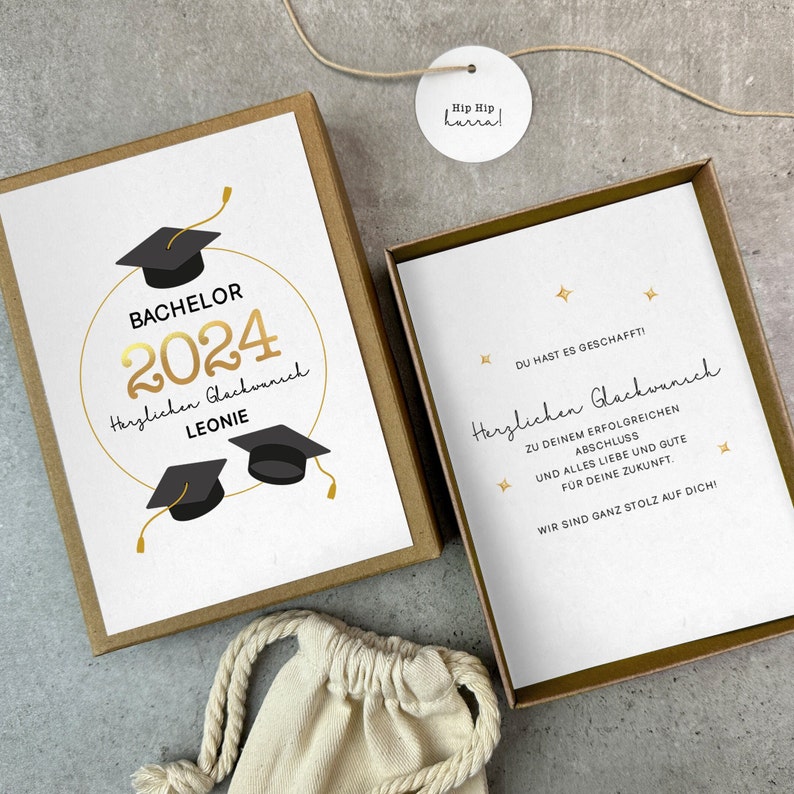 K&ouml;nnte beinhalten: Eine wei&szlig;e Karte mit goldener Schrift in einer braunen Box. Die Karte sagt "Bachelor 2024 Herzlichen Gl&uuml;ckwunsch Leonie". Es befinden sich drei schwarze Abschlusskappen auf der Karte. Die Box hat ein kleines wei&szlig;es Etikett mit der Aufschrift "Hip Hip Hurra!"