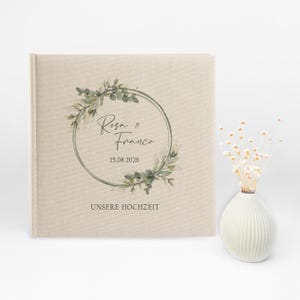 Puede incluir: Un &aacute;lbum de boda de lino beige con una corona de hojas verdes y el texto "Rosa & Franco 15.08.2026 UNSERE HOCHZEIT"