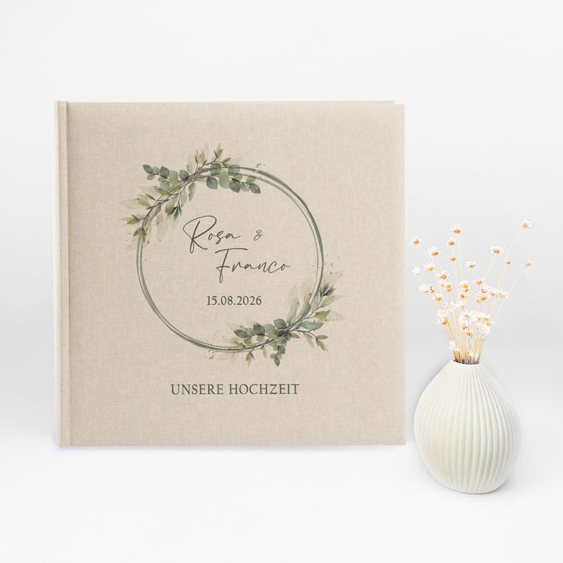 Puede incluir: Un &aacute;lbum de boda de lino beige con una corona de hojas verdes y el texto "Rosa & Franco 15.08.2026 UNSERE HOCHZEIT"