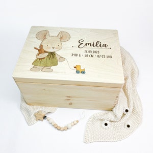 Puede incluir: Una caja de madera para recuerdos con un acabado de madera clara. La tapa presenta una ilustración pintada a mano de un ratón que sostiene un animal de peluche y tira de un pato de juguete. La caja está grabada con el nombre "Emilia" y el texto "17.09.2023 3410 g - 50 cm - 07:23 Uhr".