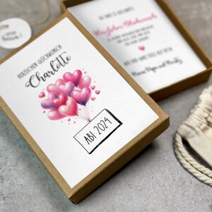 May include: A brown box with a white card inside. The card has a watercolor illustration of pink and purple heart-shaped balloons with the text "Herzlichen Gl&uuml;ckwunsch Charlotte" and "ABI 2024".