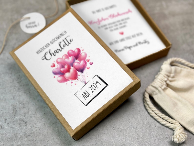 May include: A brown box with a white card inside. The card has a watercolor illustration of pink and purple heart-shaped balloons with the text "Herzlichen Gl&uuml;ckwunsch Charlotte" and "ABI 2024".