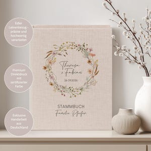 Puede incluir: Un libro beige cubierto de lino con un diseño de corona floral, los nombres "Theresa & Fabian" y la fecha "26.09.2026". La palabra "STAMMBUCH" y el apellido "Familie Pfeifer" también son visibles. El libro está sobre una superficie blanca.