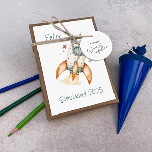 Może przedstawiać: Zestaw prezentowy na pierwszy dzień szkoły dziecka. Zestaw zawiera kartę z ilustracją rakiety, imieniem "Felix" i tekstem "Schulkind 2025". W zestawie znajduje się r&oacute;wnież niebieska torba w kształcie stożka i kredki.