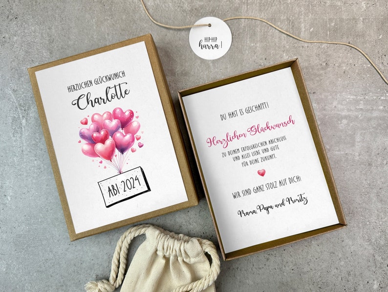 May include: A brown gift box with a white card inside. The card has a pink and purple watercolor illustration of a bunch of heart-shaped balloons with the text "HERZLICHEN GL&Uuml;CKWUNSCH Charlotte" and "ABI 2024". The card in the box has the text "DU HAST ES GESCHAFTTI! Herzlichen Gl&uuml;ckwunsch" and "WIR SIND GANZ STOLZ AUF DICH! Mama Papa und Moritz".