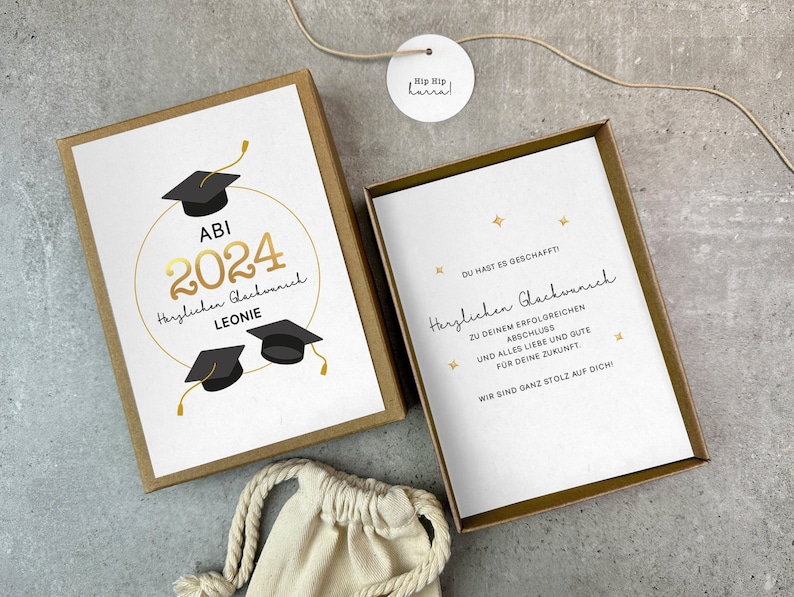 K&ouml;nnte beinhalten: Eine Karte zum Abitur mit goldenen Lettern, auf der "ABI 2024" und "Herzlichen Gl&uuml;ckwunsch Leonie" steht. Die Karte befindet sich in einer braunen Schachtel mit einer wei&szlig;en Tasche.