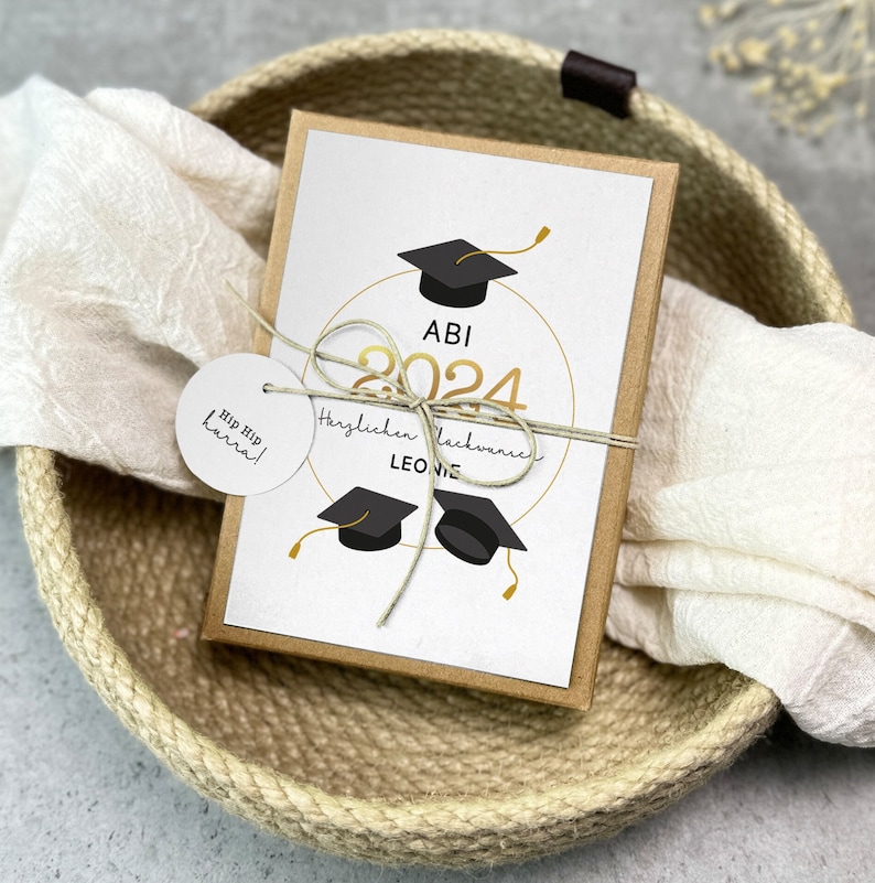 K&ouml;nnte beinhalten: Eine wei&szlig;e Karte mit goldener Schrift und einem schwarzen Abschluss-Hut-Design. Die Karte sagt "Abi 2024 Herzlichen Gl&uuml;ckwunsch Leonie". Die Karte befindet sich in einer braunen Papiergeschenkbox und ein wei&szlig;es Tuch liegt auf der Box. Die Box befindet sich in einem geflochtenen Korb.