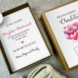 May include: A white card with a pink heart and the text "Herzlichen Gl&uuml;ckwunsch" and "Du hast es geschafft!" in a brown box. The card is decorated with pink and purple heart-shaped balloons and the text "Charlotte" and "ABI 2024".