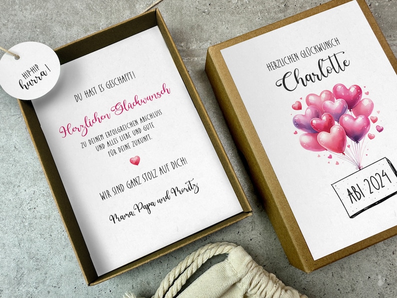 May include: A white card with a pink heart and the text "Herzlichen Gl&uuml;ckwunsch" and "Du hast es geschafft!" in a brown box. The card is decorated with pink and purple heart-shaped balloons and the text "Charlotte" and "ABI 2024".
