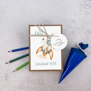 Może przedstawiać: Pudełko prezentowe z ilustracją rakiety, imieniem "Felix" i tekstem "Schulkind 2025". Niebieski stożkowy upominek na przyjęcie i trzy kredki r&oacute;wnież są obecne. Pudełko jest związane sznurkiem.