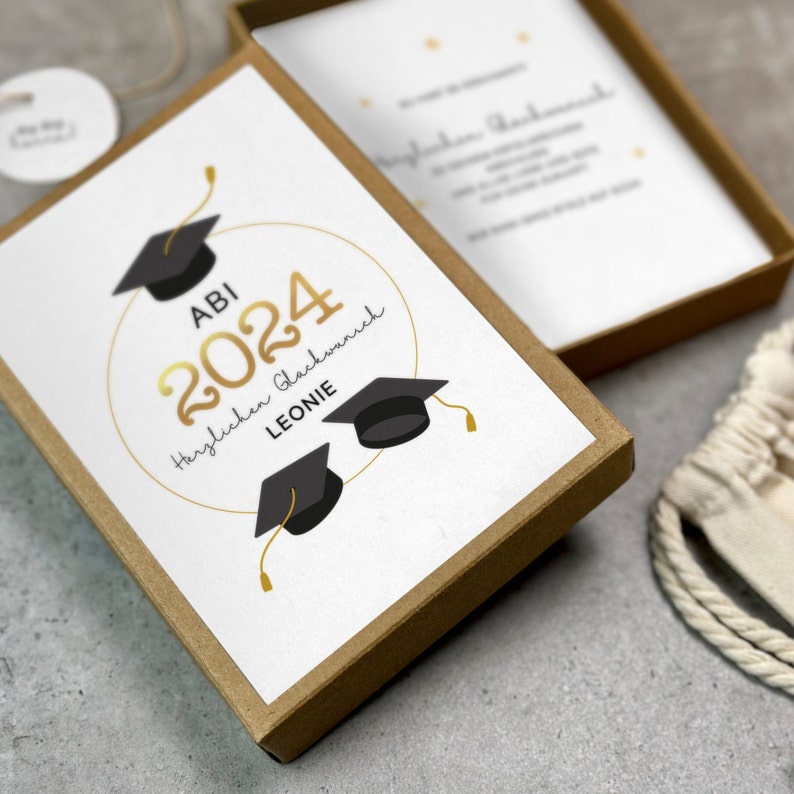 K&ouml;nnte beinhalten: Eine wei&szlig;e Karte mit goldener Schrift und schwarzen Abschlusskappen. Die Karte sagt "ABI 2024 Herzlichen Gl&uuml;ckwunsch LEONIE".
