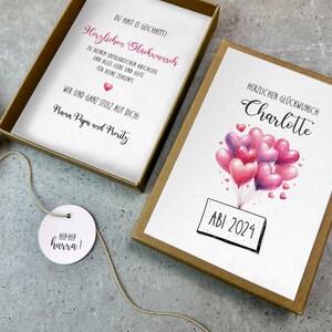 May include: A brown box with a white card inside. The card has a pink and purple heart balloon illustration with the text "HERZLICHEN GL&Uuml;CKWUNSCH Charlotte ABI 2024". The box also contains a small white bag with a tag that says "HIPHIP hurra!"