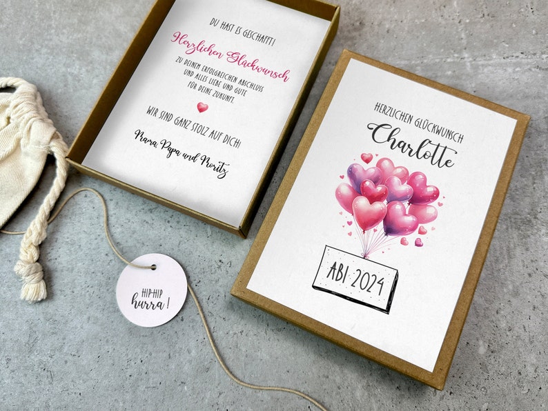 May include: A brown box with a white card inside. The card has a pink and purple heart balloon illustration with the text "HERZLICHEN GL&Uuml;CKWUNSCH Charlotte ABI 2024". The box also contains a small white bag with a tag that says "HIPHIP hurra!"