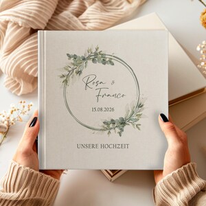 Puede incluir: Un &aacute;lbum de boda blanco con un dise&ntilde;o de corona de hojas verdes. El &aacute;lbum tiene el texto "Rosa & Franco" y la fecha "15.08.2026" impresa en la portada. El texto "Unsere Hochzeit" est&aacute; impreso debajo de la corona.