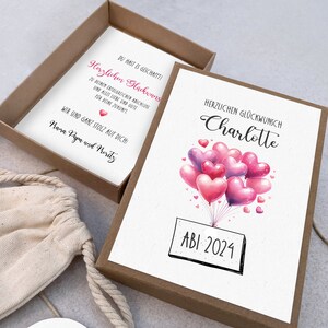 May include: A white card with pink and purple heart-shaped balloons and the text "HERZLICHEN GL&Uuml;CKWUNSCH Charlotte ABI 2024" in black. The card is in a brown box with a white card inside that says "DU HAST ES GESCHAFFTI Herzlichen Gl&uuml;ckwuns ZU DEINEM ERFOLGREICHEN ABSCHLUSS UND ALLES LIEBE UND GUTE F&Uuml;R DEINE ZUKUNFT WIR SIND GANZ STOLZ AUF DICH Mara Papa und Moritz"