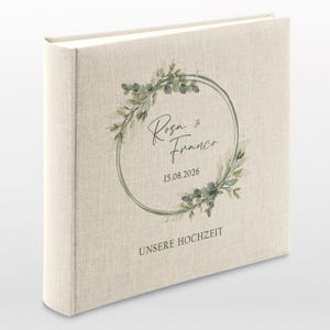 Puede incluir: Un &aacute;lbum de fotos cubierto de lino con un dise&ntilde;o de corona floral verde. El &aacute;lbum tiene el texto "Rosa & Franco" y "15.08.2026" en letra cursiva negra. El texto "UNSERE HOCHZEIT" est&aacute; impreso en negro en la parte inferior del &aacute;lbum.